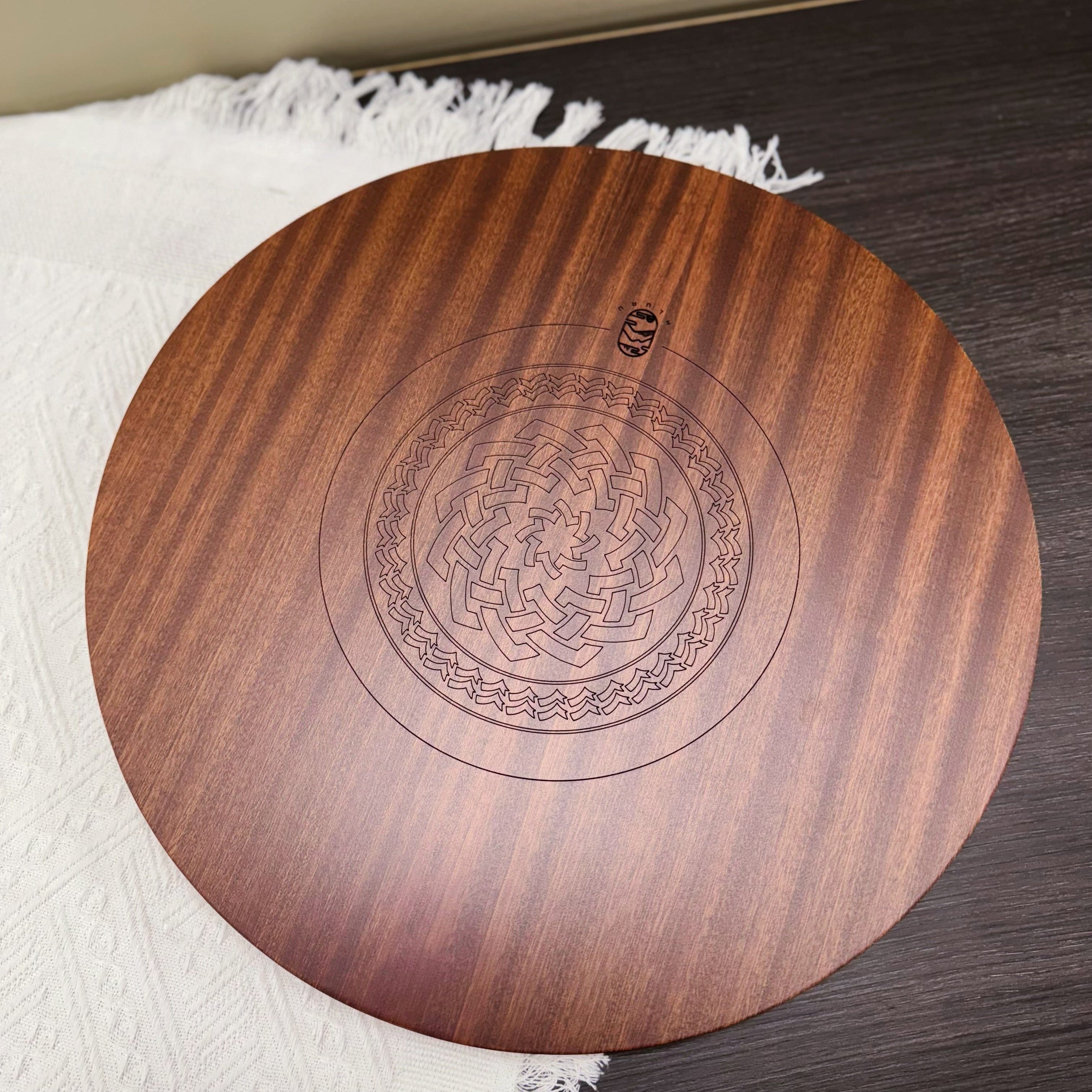 Round wooden tongue drum with engraved mandala design, resting on white fringed cloth