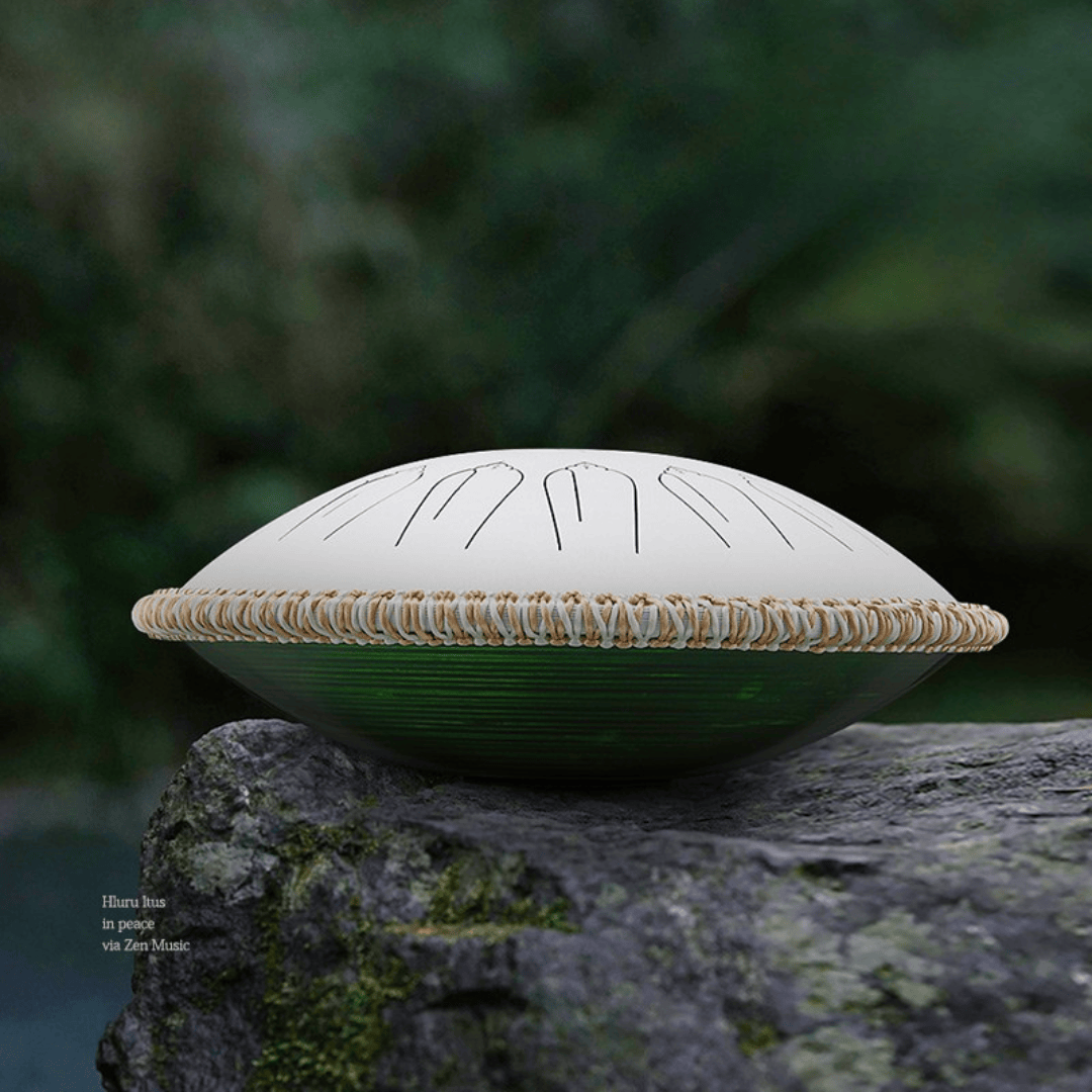 White Hluru EQ Titanium Steel Tongue Drum 15 Notes 14'' with rattan surround and wooden base, shown outdoors on a rock.