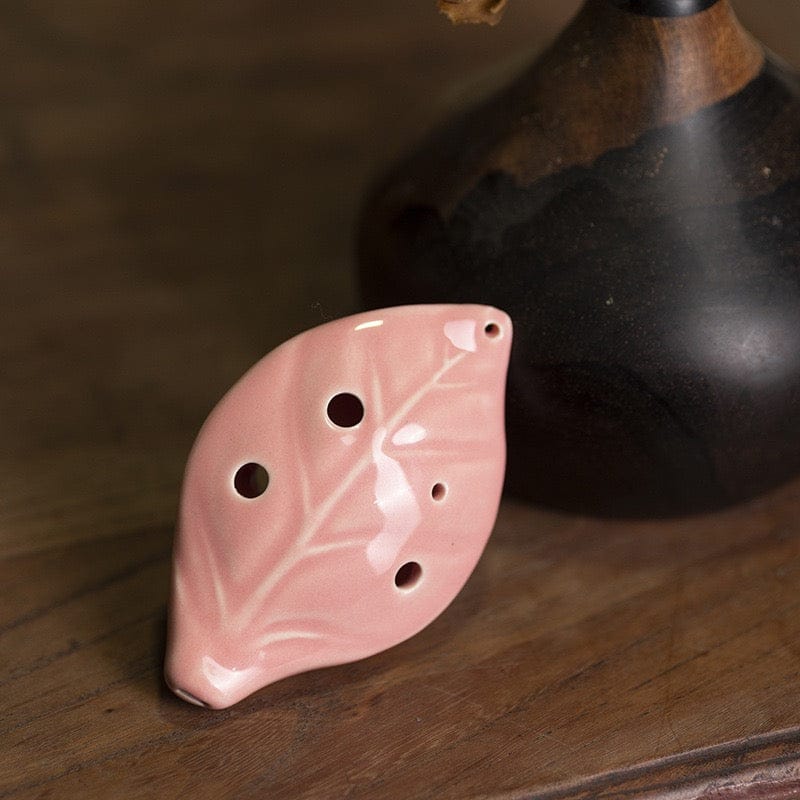Pink leaf-shaped ocarina with delicate vein details, resting on wooden surface beside dark vase