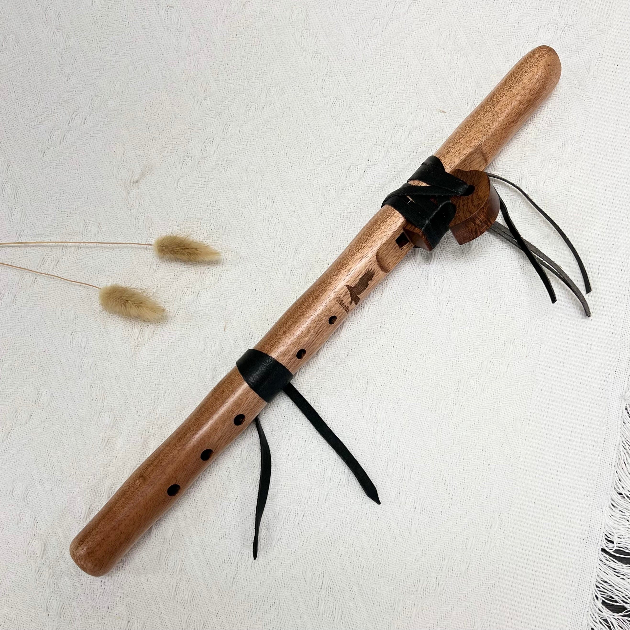 Handcrafted wooden flute with leather ties and eagle engraving, resting on textured white fabric with dried grass accents.