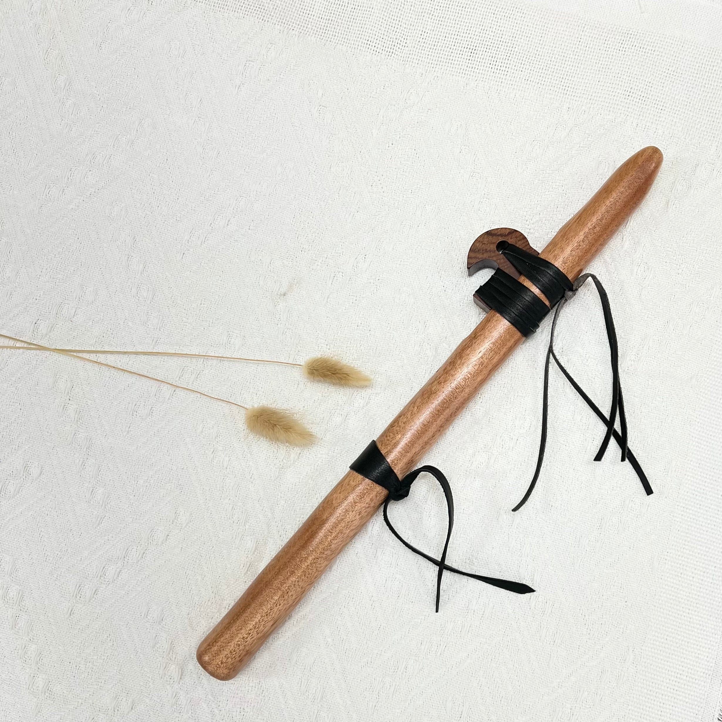 Handcrafted wooden tongue drum mallet with black leather straps, resting on white fabric with dried grass accents