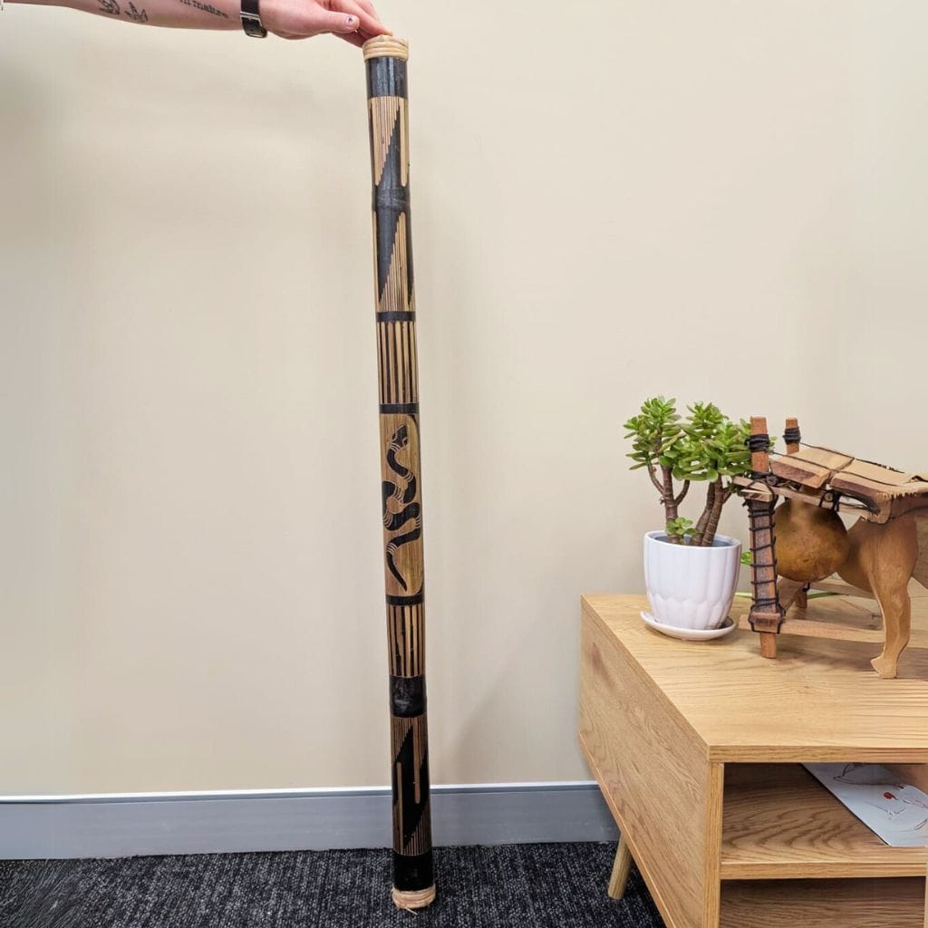 Hand holding a decorated didgeridoo with tribal patterns, next to a wooden side table with a plant and kalimba.