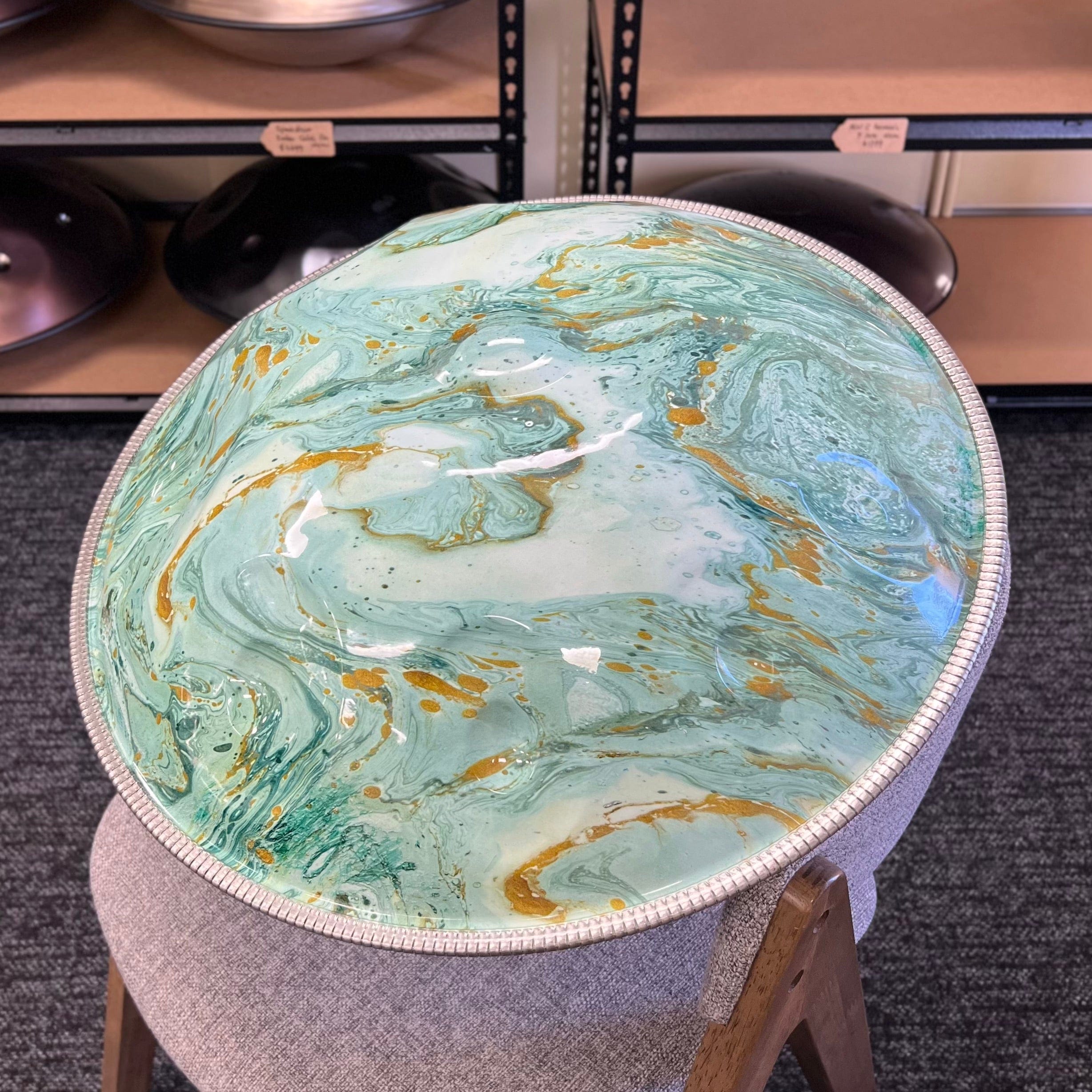 Handpan with swirling teal, gold, and white resin art, displayed on stool in music store