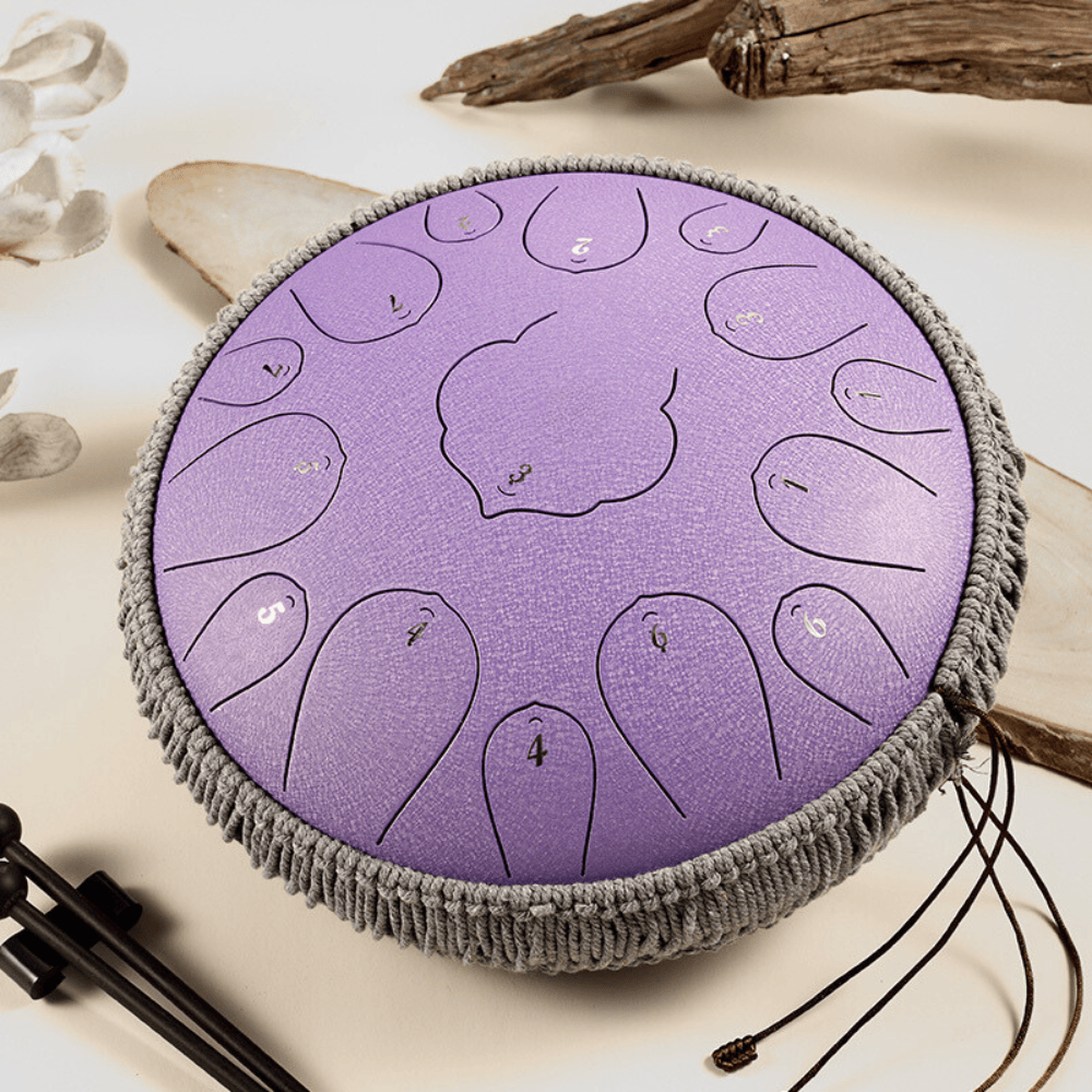 Purple handpan drum with numbered tone fields, grey fringe, and mallets, styled with driftwood and petals