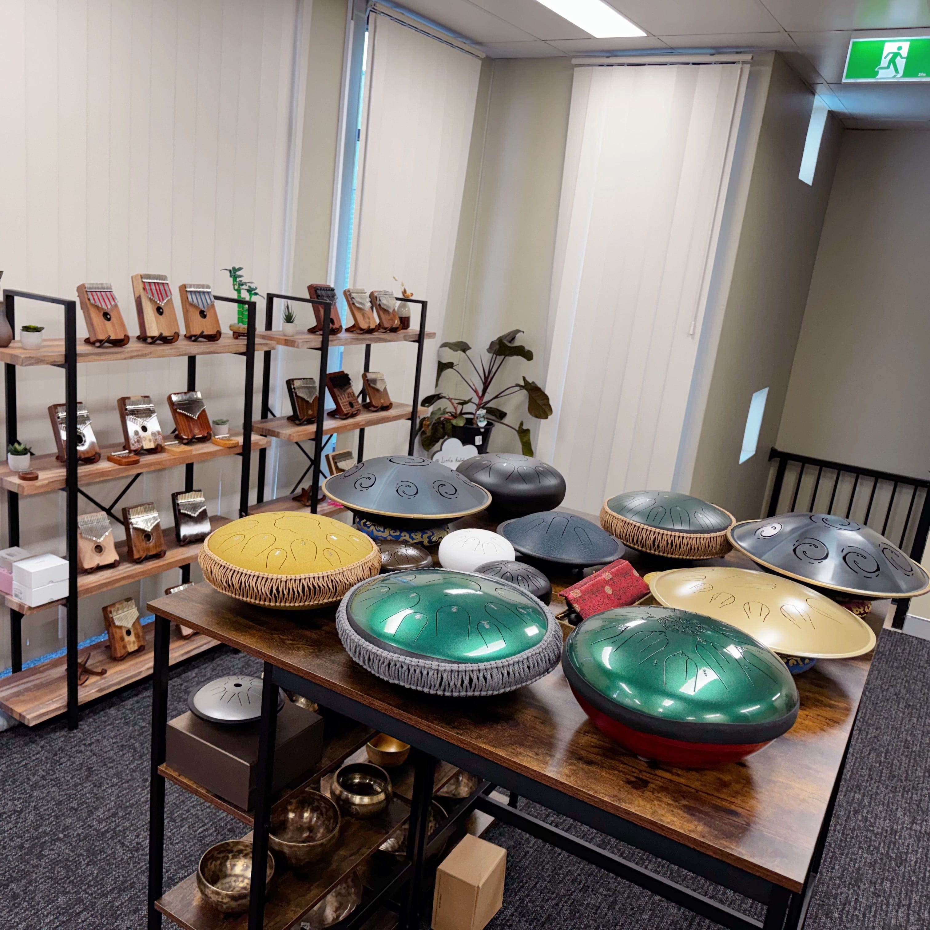 A collection of handpan and steel pan instruments displayed on a wooden table in an indoor showroom or studio, with shelves of other musical instruments in the background.