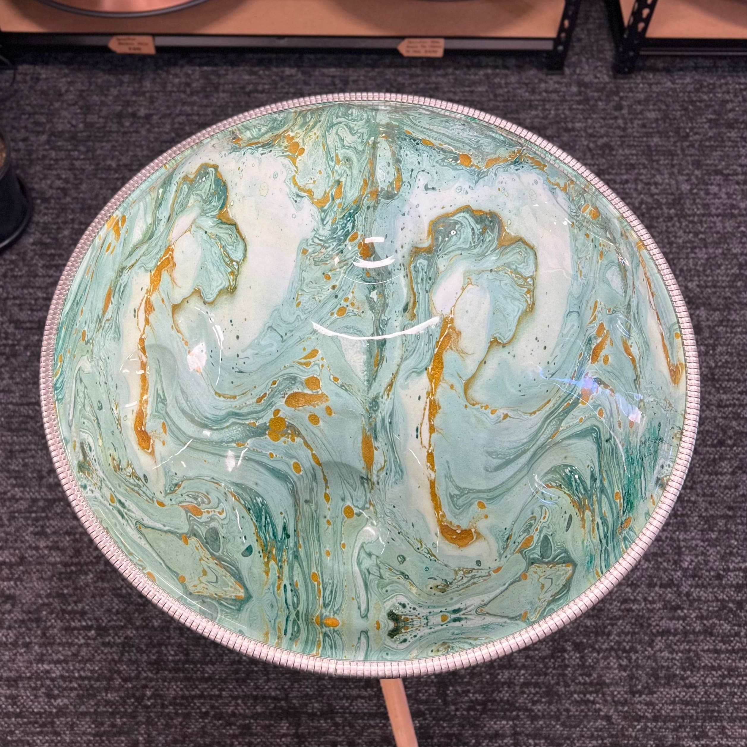 Handpan with swirling teal, white, and gold resin art, displayed on stand in store